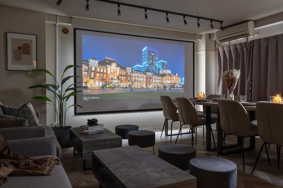 プロジェクターに東京駅の夜景が映し出された室内。間接照明のみの落ち着いた空間で、ソファとダイニングテーブルがスクリーンを囲むように配置されている。