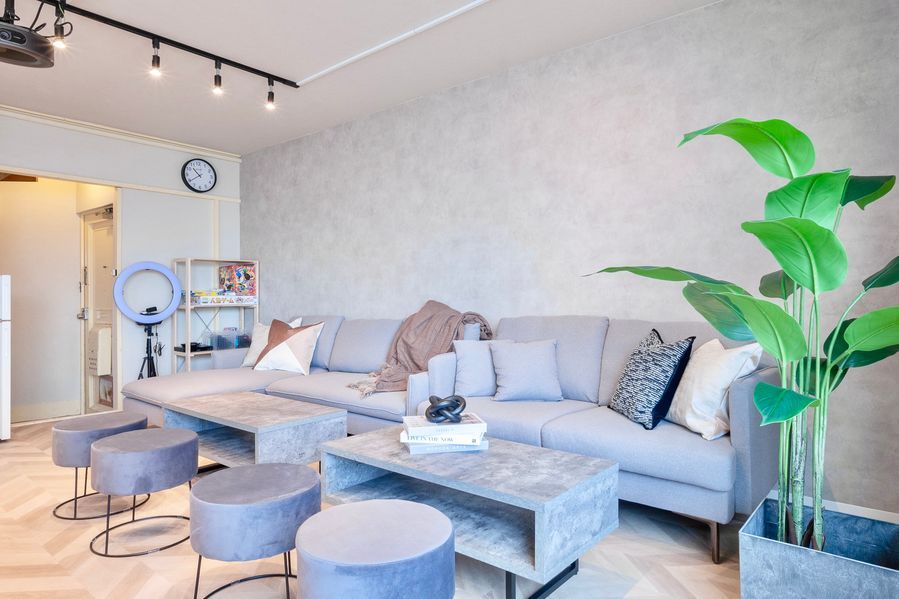 ソファエリアの全景。コンクリート調の壁面、L字ソファ、グレーのベルベットスツール3脚、リングライトの三脚が奥に見える。入口付近にはボードゲーム棚。